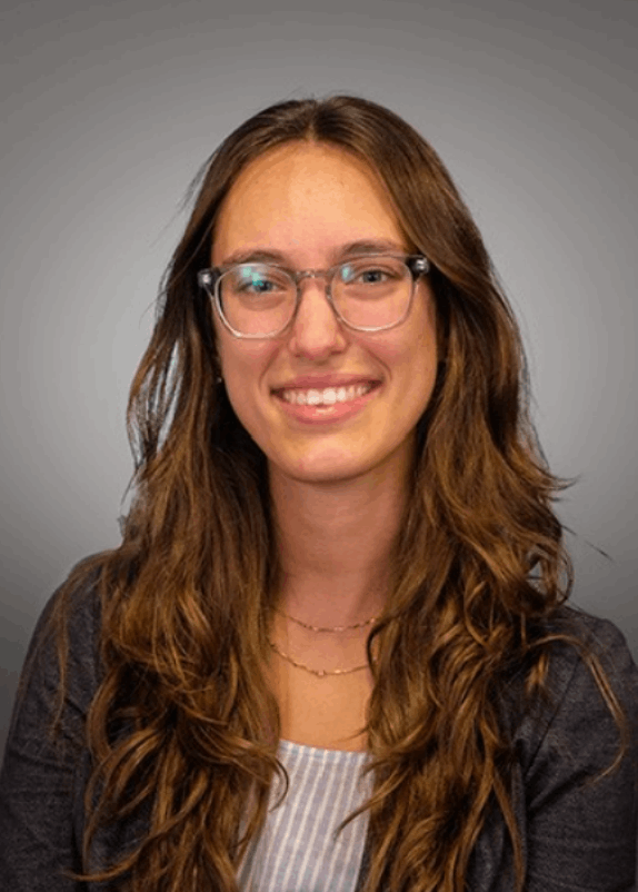 Smiling woman with long wavy brown hair and clear glasses wearing a dark blazer over a striped top.