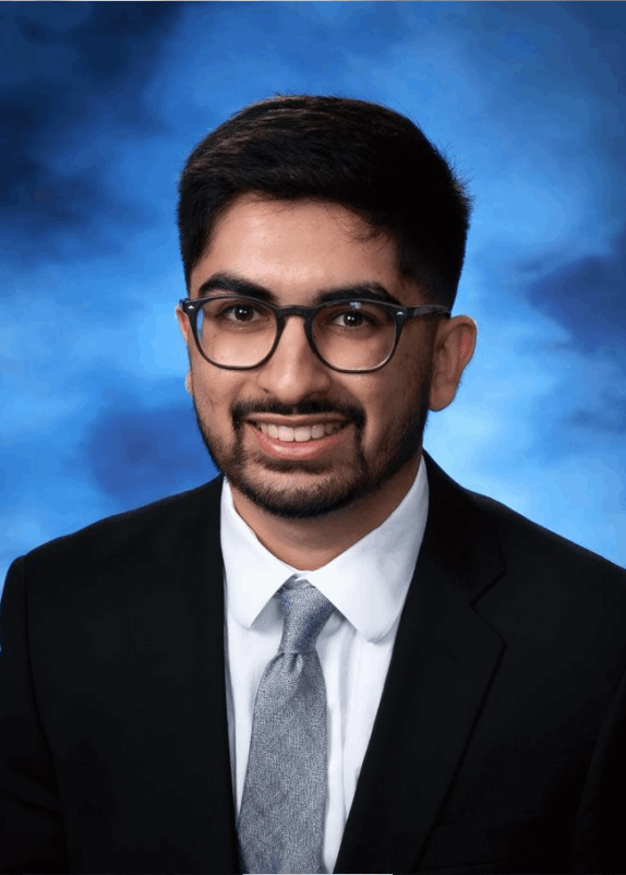 Smiling man with dark hair, beard, and glasses wearing a black suit and blue tie against a blue backdrop.