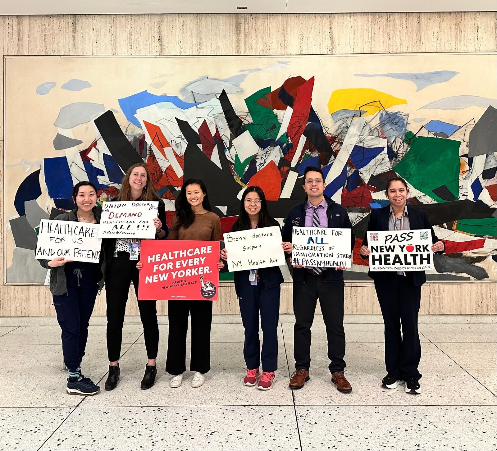 Six people stand in a row holding signs advocating for healthcare reform in New York, positioned in front of a large, colorful abstract mural wall.