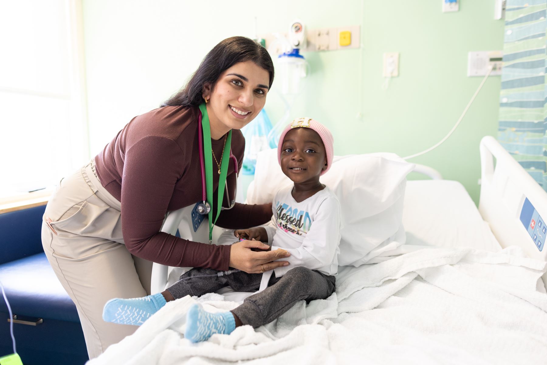 A smiling female doctor with a stethoscope leans over a young child in a hospital bed, who is wearing a pink beanie and a white long-sleeve shirt.