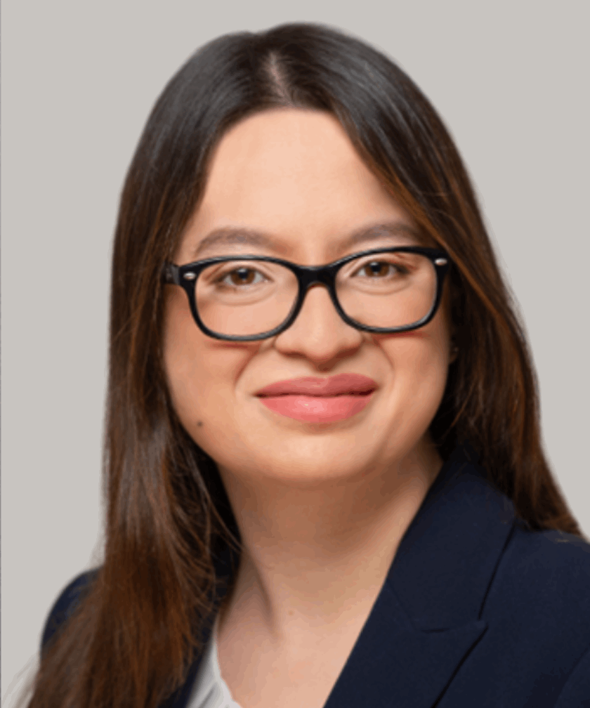 Smiling woman with long dark hair and black glasses wearing a navy blazer and white top.