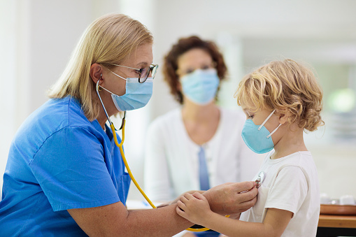 A doctor wearing a mask performs a check-up on a young child using a stethoscope.